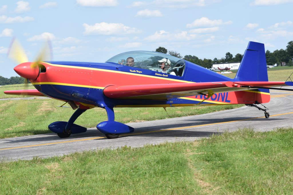 An Extra 300L in red, blue and yellow competition livery taxiing on a taxiway, two people in the cockpit, the canopy rail lettered Neville Hogan and Sheldon Apsell.